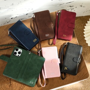 May include: Six different coloured phone cases with a wallet flap and a wrist strap. The cases are made of faux leather and have different initials on them. The cases are arranged on a wooden surface.