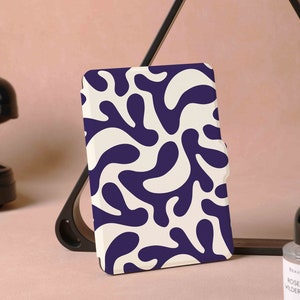 Puede incluir: Una funda de teléfono con un fondo crema y un patrón repetitivo de formas orgánicas azul oscuro. La funda está apoyada en un soporte negro, con un teléfono negro vintage y un frasco de perfume en el fondo.