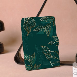 Puede incluir: Una funda para lector electrónico de color verde y dorado con un diseño floral. La funda tiene un contorno dorado y un fondo verde con hojas doradas.