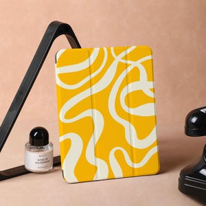 May include: A yellow and white tablet case with an abstract design. The case is on a black triangular stand. A small bottle of perfume is in front of the stand. A black rotary phone is on the right side of the image.