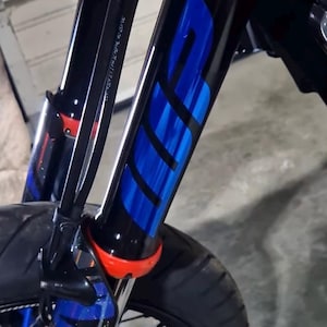 Puede incluir: Primer plano de la horquilla de suspensión delantera de una motocicleta. La horquilla es negra con gráficos azules y negros. La horquilla tiene un componente rojo cerca del neumático. El neumático es negro y la rueda tiene radios azules.