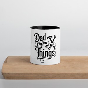 Puede incluir: Taza de cerámica blanca con borde y asa negros. La taza tiene un gráfico negro que dice "Dad Fixer of Things" con un diseño de llave inglesa y martillo.