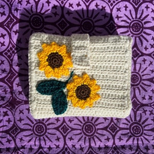 May include: A crocheted cream-colored pouch with two embroidered sunflowers. The sunflowers have brown centers and yellow petals. The pouch is on a purple fabric background with a repeating pattern.