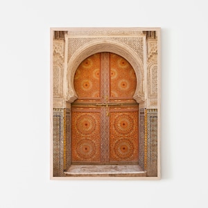 Puede incluir: Una impresión enmarcada que presenta una vista detallada de una puerta arqueada ornamentada. La puerta de madera es de color naranja cálido con intrincados patrones circulares. El arco y las paredes circundantes están adornados con tallas detalladas y mosaicos.