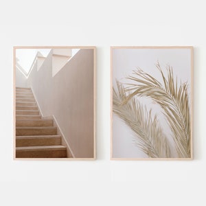 Op de afbeelding: Twee ingelijste prints, een met een beige trap die naar een witte muur met een raam leidt, en de andere met een close-up van een gedroogde palmblad tegen een witte achtergrond.