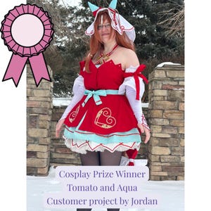May include: A red and aqua cosplay costume with gold detailing, including a heart motif on the skirt. The outfit is complemented by a matching headpiece and is labelled "Cosplay Prize Winner".