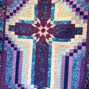 Peut inclure: Une courtepointe avec un motif de croix. La croix est composée de tissus violets, bleus et blancs. Le motif géométrique comprend une étoile au centre. La bordure est ornée de motifs floraux.