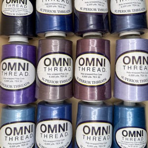 May include: A collection of twelve spools of Omni Thread poly-wrapped poly core thread in various shades of blue and purple. Each spool features a white label with the text Omni Thread, Superior Threads, 6,000 yds, and TEX 30.