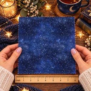 May include: A square fabric swatch featuring a dark blue night sky pattern with white stars and nebulae, held above a wooden ruler that measures 8 inches in length, with pinked edges along the perimeter of the fabric piece.