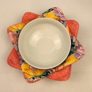 May include: A white ceramic bowl sits on top of two floral patterned pot holders. The pot holders are pink with a floral design in shades of yellow, purple, and green.