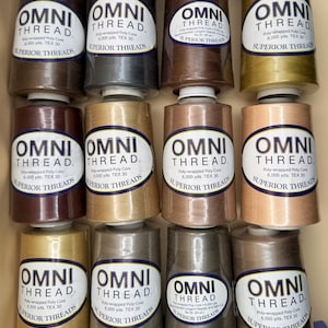 May include: A collection of twelve large spools of Omni Thread by Superior Threads, arranged in a grid. Each spool features a white oval label with black text, indicating poly-wrapped poly core, 6,000 yards, and TEX 30. The threads are in various neutral shades including brown, tan, gray, and gold.