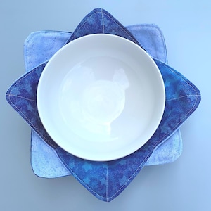 May include: A white bowl sits inside a blue and white fabric bowl cozy with a star pattern. The cozy is made of two layers of fabric.