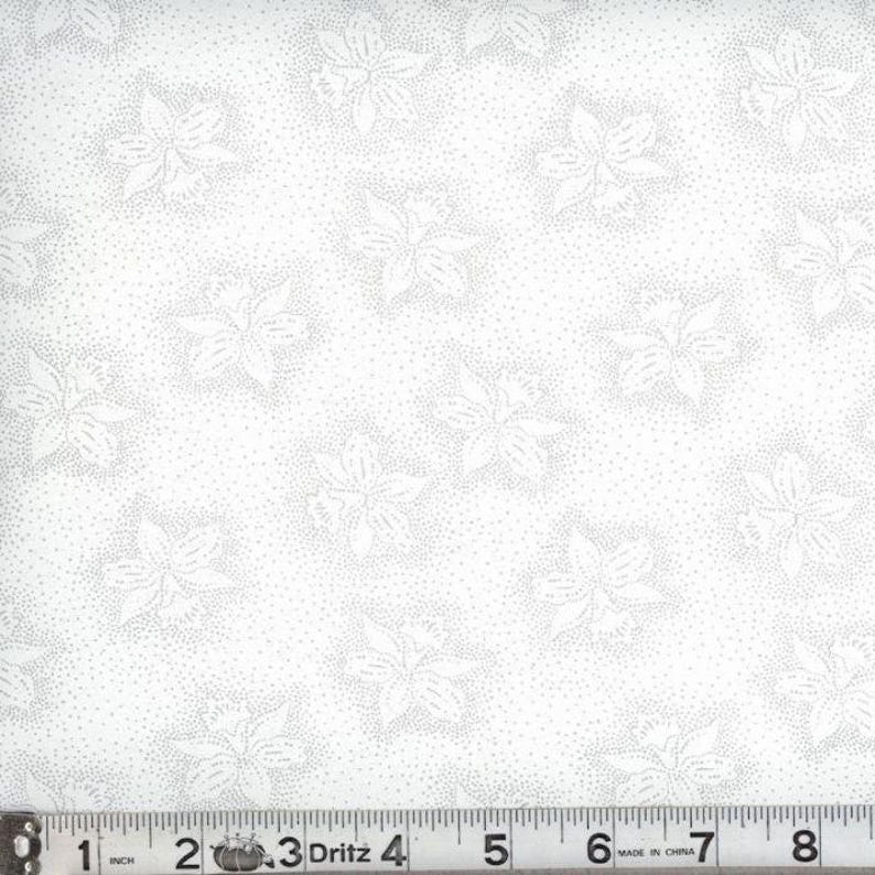 May include: A white fabric featuring a repeating pattern of light gray floral outlines surrounded by fine stippled dots. A Dritz measuring tape is positioned at the bottom, showing a scale of 8 inches across the width of the textile material.