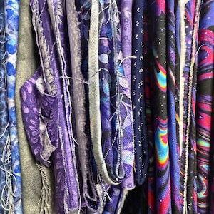 May include: Close-up of a collection of fabric swatches in shades of blue, purple, and gray. The fabrics feature various patterns, including floral designs and abstract prints. Some swatches have frayed edges and visible stitching.