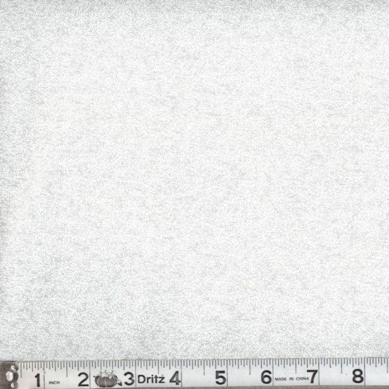 May include: A light gray fabric featuring a dense, repeating white pattern of small, delicate leaves and vines. A Dritz measuring tape is positioned along the bottom edge, showing a scale in inches that extends past 8 inches in total length.