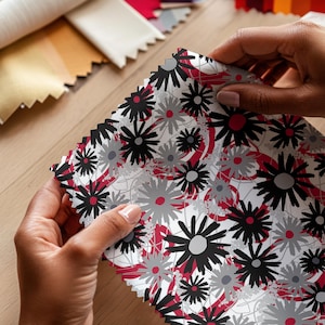 May include: A square fabric swatch featuring a repeating floral pattern with black and gray daisy-like flowers on a white background, accented by thin red circular lines and abstract red shapes throughout the design with pinked edges.