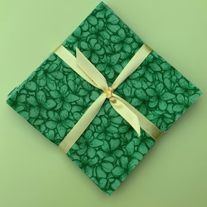 May include: A square stack of fabric squares featuring a repeating dark green floral pattern on a lighter green background. The stack is tied diagonally with a pale yellow satin ribbon that is knotted in the center of the top fabric square.