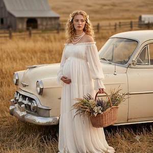 May include: A floor-length off-the-shoulder white gown with long sleeves and a tiered skirt. The dress features a gathered bodice and a flowing silhouette. A woven wicker basket filled with dried flowers is held in one hand.