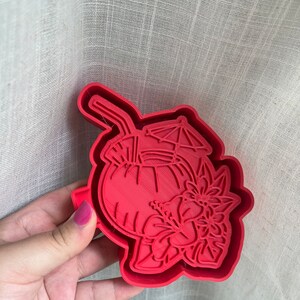 Peut inclure: Emporte-pièce en plastique rouge en forme de boisson tropicale avec une paille, un parapluie et une fleur d'hibiscus.
