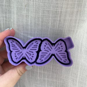 May include: Purple plastic cookie cutter shaped like two butterflies. The butterflies are facing each other with their wings spread out.
