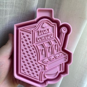 May include: Pink plastic cookie cutter shaped like a slot machine with the words "Lucky Cowgirl" on the front. The slot machine has three windows with the number 7 in each window.