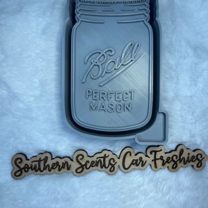 May include: A gray plastic mold of a Ball mason jar with the text "PERFECT MASON" on the front. The mold is sitting on a white fluffy surface. Below the mold is a wooden sign that reads "Southern Scents Car Freshies".