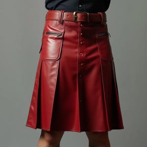 Op de afbeelding: Een rode leren kilt met bijpassende riem en gesp. De kilt heeft twee zakken met ritssluiting en decoratieve knopen aan de voorkant. Het geplooide ontwerp voegt structuur toe aan het kledingstuk.