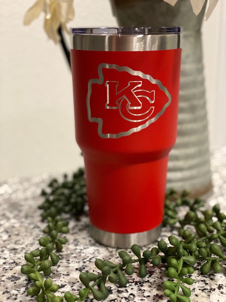 May include: A red stainless steel tumbler with a silver lid. The tumbler has a white, engraved design of a Native American arrowhead with the letters "KC" inside.
