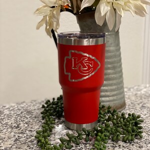 May include: Red stainless steel tumbler with a silver lid and a silver base. The tumbler has a white and silver logo of "KC" with an arrowhead on either side of the letters.