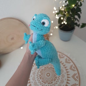 May include: A crocheted turquoise blue dinosaur toy with white eyes and a pink tongue. The dinosaur is being held in a hand.