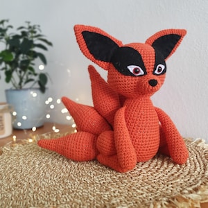 May include: A crocheted orange fox with black ears and eyes. The fox has a curled tail and is sitting on a woven straw mat.