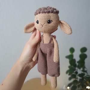 Peut inclure: Un animal en peluche crocheté en forme de mouton portant une salopette brune. Le mouton a une tête brune et un corps brun clair. Le mouton a des yeux noirs et un petit nez rose.