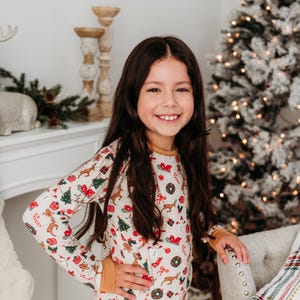 May include: A young girl wearing a white long-sleeved pajama top with a festive Christmas print. The print features reindeer, Christmas trees, presents, candy canes, and other holiday symbols. The pajamas have a yellow trim around the cuffs and neckline.