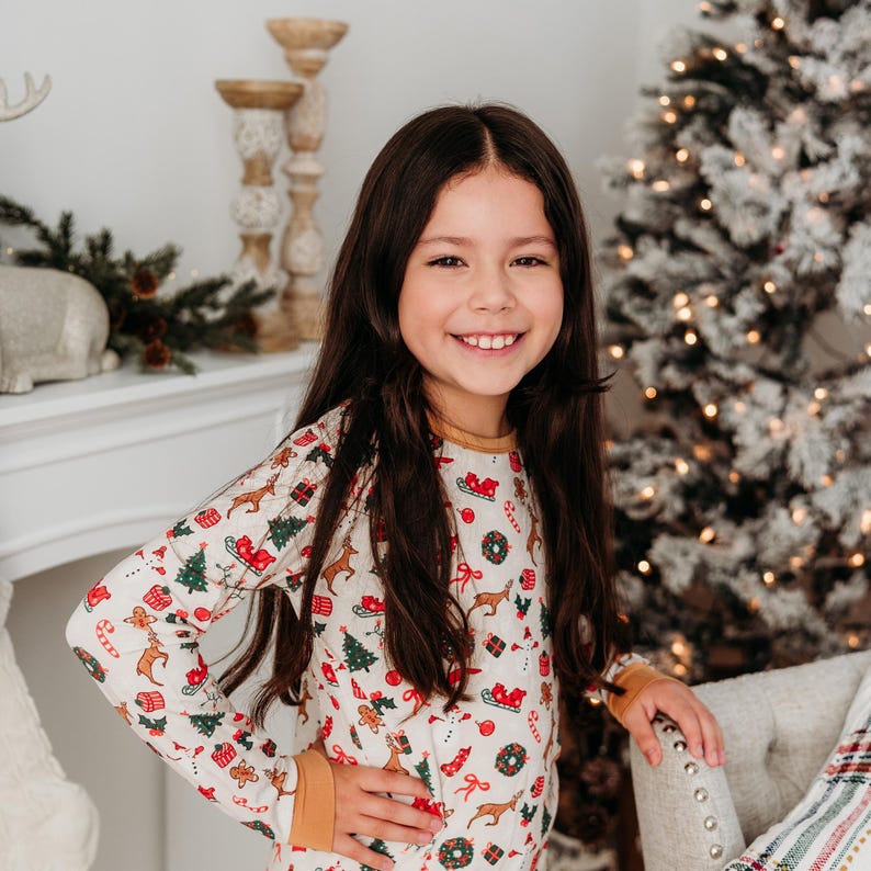 May include: A young girl wearing a white long-sleeved pajama top with a festive Christmas print. The print features reindeer, Christmas trees, presents, candy canes, and other holiday symbols. The pajamas have a yellow trim around the cuffs and neckline.