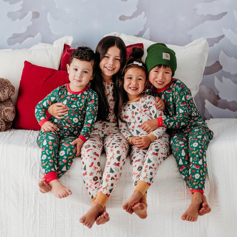 May include: Four children wearing matching Christmas pajamas with a green and white pattern. The pajamas feature a variety of holiday-themed designs, including candy canes, ornaments, and reindeer. The children are sitting on a white couch with a red pillow behind them. One child is wearing a green hat.
