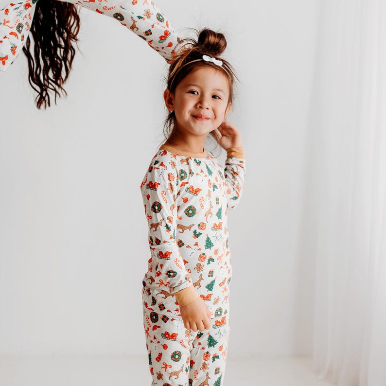 May include: A young girl wearing a white long-sleeved pajamas with a Christmas print. The pajamas feature a variety of festive designs, including reindeer, candy canes, and ornaments. The girl is smiling and looking at the camera.