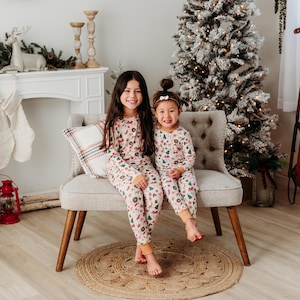 May include: Two children wearing matching white pajamas with a Christmas-themed print. The pajamas feature a red, green, and gold pattern with images of reindeer, snowmen, and Christmas trees. The children are sitting on a white couch in front of a Christmas tree.