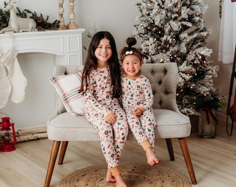 Puede incluir: Dos niños vistiendo pijamas blancos a juego con un estampado navideño. Las pijamas tienen un patrón rojo, verde y dorado con imágenes de renos, muñecos de nieve y árboles de Navidad. Los niños están sentados en un sofá blanco frente a un árbol de Navidad.