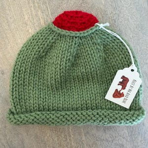 May include: A hand-knitted green beanie with a red top. The hat has a folded brim and a tag that reads "Knit by Ma and Bear" with a bear illustration. The hat is made of a soft, textured yarn.