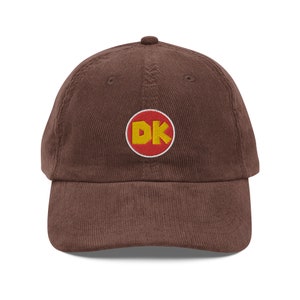 Peut inclure: Une casquette de baseball en velours côtelé marron avec un cercle rouge et les lettres "DK" en jaune.