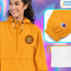 Puede incluir: Una chaqueta amarilla con capucha, cierre de cremallera y un parche con el logotipo de una corona en el pecho izquierdo. La chaqueta tiene capucha con cordón y puños elásticos. La chaqueta se muestra con una bolsa de cordón blanca.