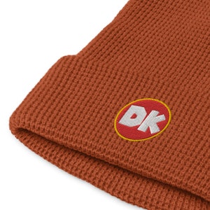 Puede incluir: Gorro de color óxido con textura de punto de gofre. El gorro presenta un parche bordado con las letras "DK" en blanco, contorneadas en amarillo, sobre un fondo rojo. El gorro está doblado en el borde.
