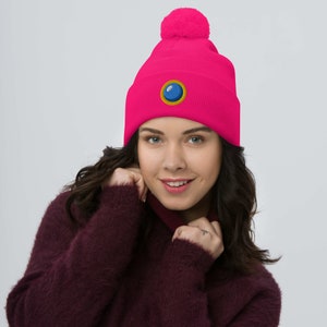 Puede incluir: Un gorro rosa con un pompón en la parte superior y un diseño circular bordado en azul y dorado.