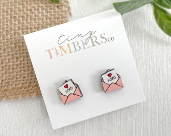 May include: A pair of pink wooden earrings shaped like envelopes with a white card inside that says "XOXO" and has a red heart. The earrings are on a white card with the text "tiny TIMBERSco".