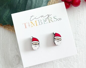 May include: A pair of wooden Santa Claus stud earrings. The earrings are red and white with a black outline. The earrings are on a white card with the text "tiny TIMBERSco" printed on it.