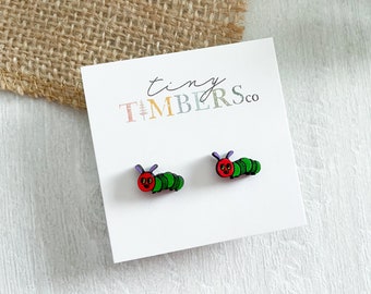 May include: A pair of wooden earrings featuring a green caterpillar with a red head and purple antennae. The earrings are displayed on a white card with the text "tiny timbers co."