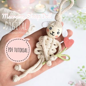 May include: A handmade macrame sheep doll with a wooden head, white ears, and a red heart. The doll is held in a hand, with the text "Macrame Sheep Doll DIGITAL PDF TUTORIAL" visible in the image.