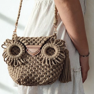 Könnte beinhalten: Beigefarbene Makramee-Handtasche in Eulenform mit langem Schulterriemen. Die Tasche hat kreisförmige Augendetails, einen Metallverschluss und einen Quastenanhänger. Die Tasche ist aus geflochtenem Seilmaterial.