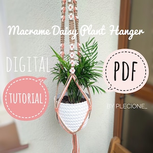 May include: Macrame Daisy Plant Hanger DIY tutorial PDF. A white pot with a green plant hangs from a macrame plant hanger with a daisy pattern. The hanger is made with natural jute twine and peach coloured yarn.