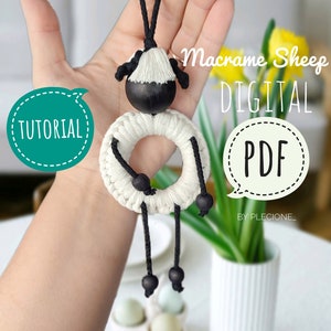 Könnte beinhalten: Ein weiß-schwarzes Makramee-Schaf-Ornament mit schwarzen Holzperlen. Das Ornament hängt an einer schwarzen Schnur und hat einen weißen und schwarzen Holzkopf. Der Text "Macrame Sheep DIGITAL PDF BY PLECIONE." ist im Bild sichtbar.